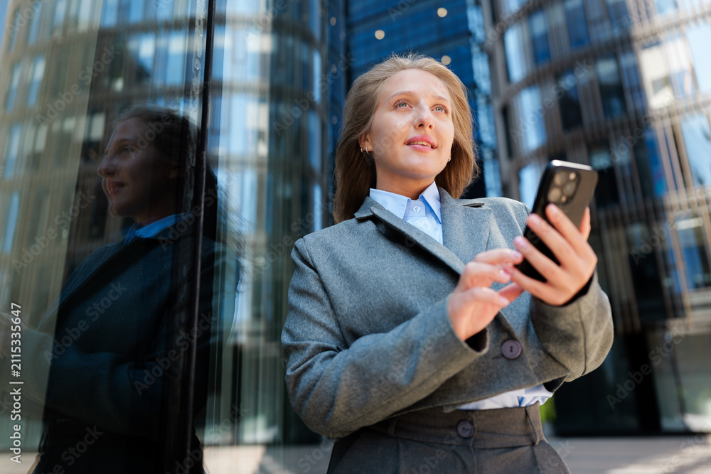 Fototapeta premium Standing confidently outside a modern building, a woman engages with her smartphone while dressed in professional attire