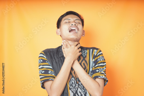 Photography Young Asian man in traditional batik shirt holding neck having sore throat or choking expression over yellow background photography