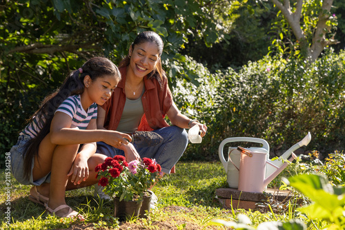 African American mom and youth daughter kneeling in yard, tending potted red blooms near water cans