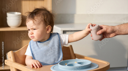 A baby sitting in a high chair looking away with a grumpy expression while being offered a sippy cup by a parents hand