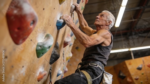 Senior male rock climber scaling indoor wall with chalked hands. Agile older athlete demonstrating strength and focus on climbing holds.