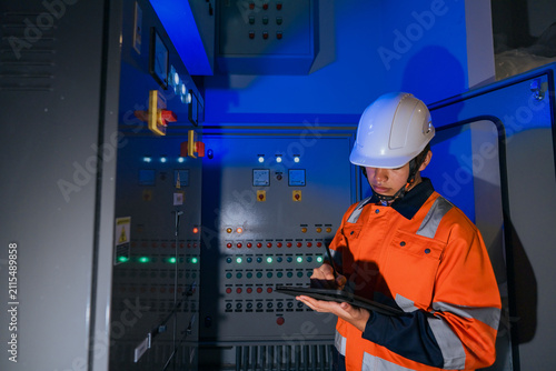 Smart Asian industrial engineer recording data on a digital tablet during electrical system evaluation at a main control panel in a modern manufacturing plant with futuristic blue lighting.