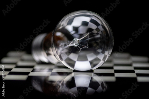 фотографія A close-up shot of a clear glass incandescent light bulb lying horizontally on a black and white checkered surface with reflections