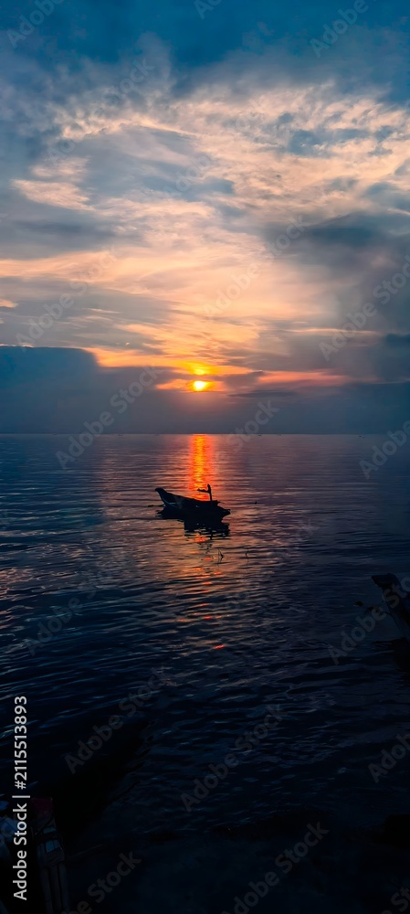 Obraz premium A powerful and dramatic seascape showing a lone fishing boat silhouette on a dark ocean beneath a dramatic sunset sky with vivid orange and red hues between dark moody storm clouds, evoking solitude.