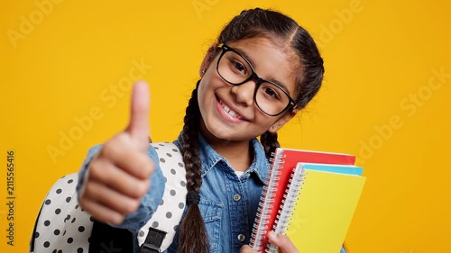 A smiling young girl with glasses giving a thumbs up while holding notebooks