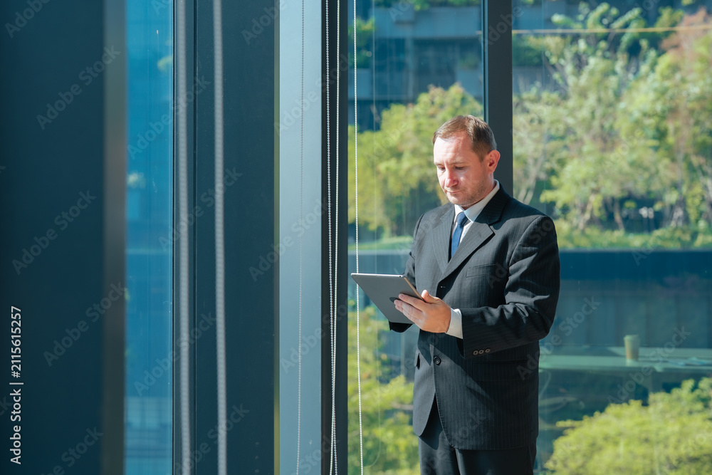 Fototapeta premium Businessman using tablet standing beside office window. Professional reviewing business information, checking reports, managing projects and working with digital technology in corporate workspace.