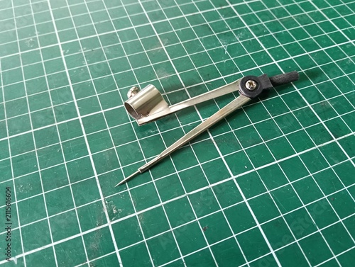 Metal drawing compass with a sharp needle point and pencil holder, used for drawing circles and arcs in technical drawing, drafting, geometry, and school work, isolated on a green cutting mat.