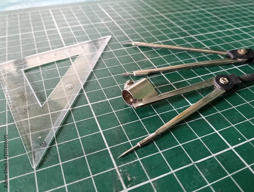 Technical drawing tools including a metal drawing compass, transparent triangular ruler, and measuring instruments arranged on a green cutting mat, used for geometry, drafting, engineering, and precis