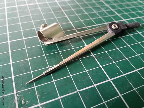 Metal drawing compass with a sharp needle point and pencil holder, used for drawing circles and arcs in technical drawing, drafting, geometry, and school work, isolated on a green cutting mat.