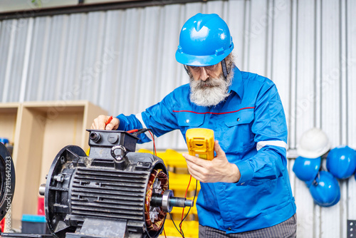 Senior industrial electrician in safety gear uses a digital multimeter to test an electric motor in a workshop. Experienced technician performing machinery maintenance and electrical diagnostics.
