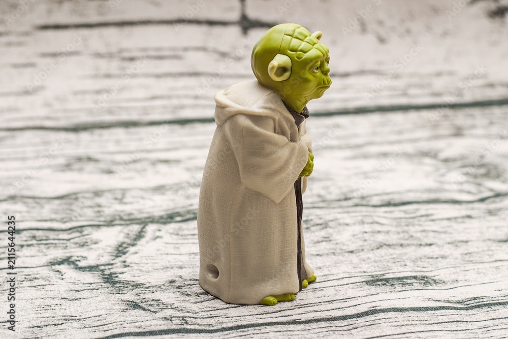 Fototapeta premium Detailed green Yoda figure stands on a textured surface, wearing a tan robe and holding a staff, exuding wisdom and tranquility