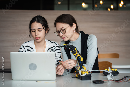 Robotics Education with an Asian female teacher guiding a young woman using a laptop beside a robotic arm during a practical technology lesson in class