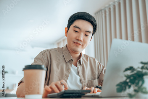 Happy young Asian man working on laptop in living room. Freelance worker male sitting at desk writing notes while watching webinar, studying online