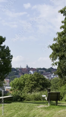 Scenic view of a green park in Flensburg, wooden bench overlooking a city skyline under a bright blue sky 