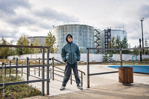 A confident teenage boy stands outdoors in front of large industrial storage tanks under dramatic cloudy skies, wearing a hooded jacket, cap, and casual streetwear