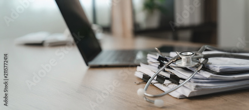Stethoscope rests on clipped paper documents beside an open laptop on a wooden desk, with wide copy space for medical administration, healthcare records, and presentation messaging.