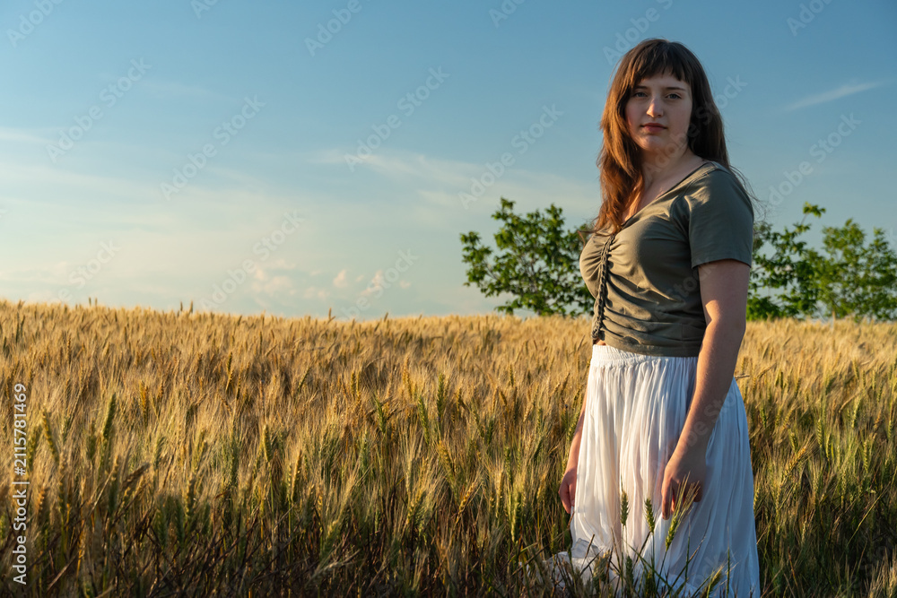 Obraz premium Young woman standing in golden wheat field at sunset