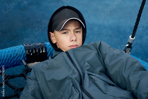 A serious teenage boy relaxes in a rope playground hammock while wearing a hooded jacket and baseball cap on an outdoor sports court. His thoughtful expression create a contemporary youth portrait