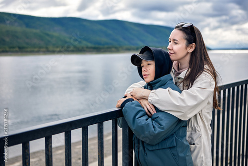 A caring mother embraces her teenage son as they quietly admire a calm mountain lake beneath dramatic cloudy skies. Dressed in warm outdoor jackets, they lean against a waterfront railing