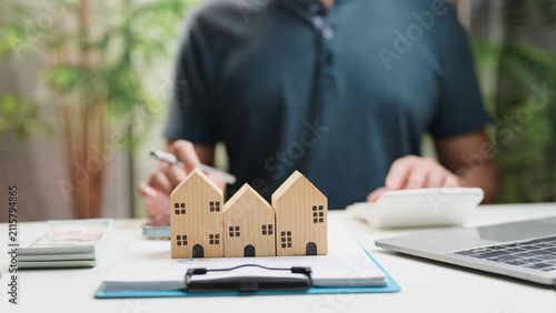 Male accountant calculates real estate investment cost sitting near wooden house model, clipboard document, calculator, laptop, money stack, planning home purchase mortgage loan.