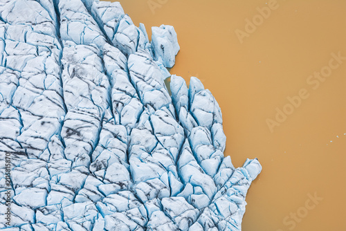Aerial view of Jokulsarlon glacier edge with deep blue crevasses and textured ice formations meeting the calm glacial lagoon water Sveitarfelagid Hornafjordur, Iceland.