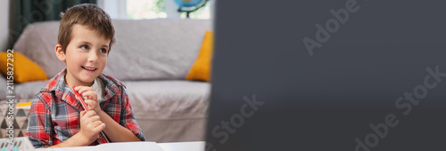 Fotografie A preschool boy learns to write capital and cursive letters in a copybook at home, sitting at a desk near a laptop