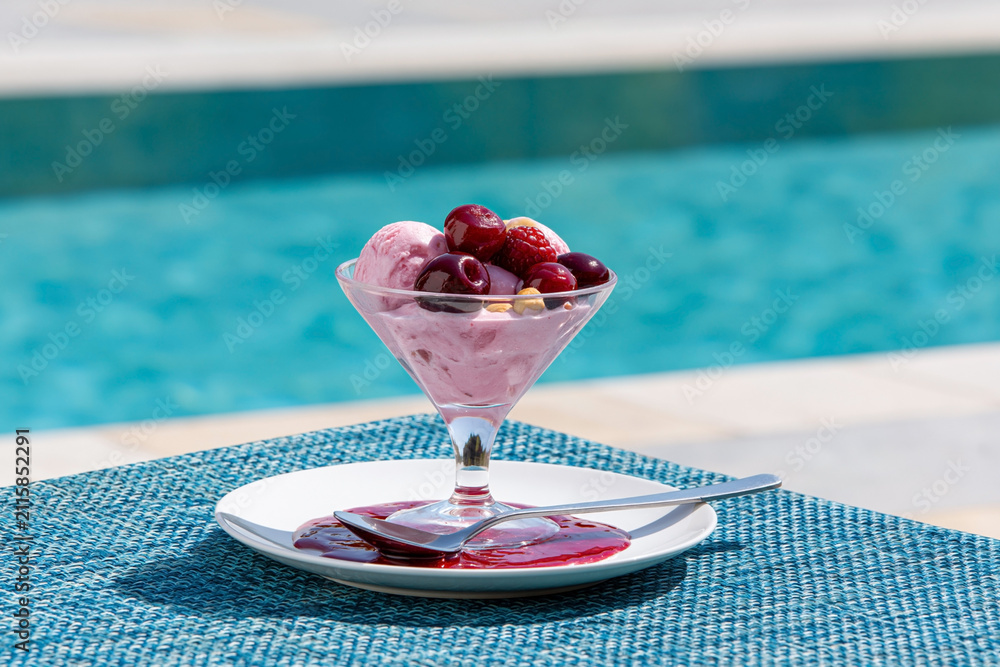 Obraz premium Berry ice cream with cherries and raspberries served in glass dessert bowl by swimming pool on sunny summer day