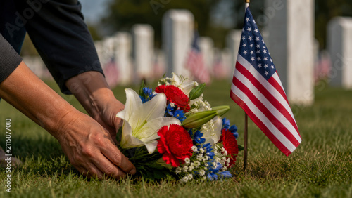 Person placing bouquet of red white and blue flowers at military grave in cemetery decorated with American flags to honor soldiers on Memorial Day or Veterans Day observance