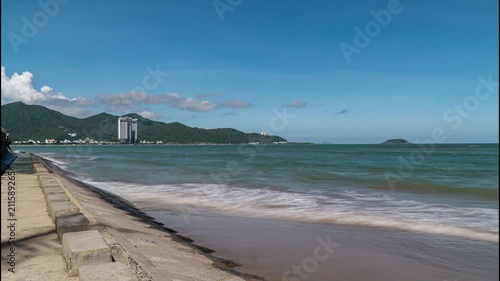 Ocean waves wash against the coastal seawall in Nha Trang, Vietnam, with green mountains, modern buildings and a tropical shoreline under a clear blue sky.
