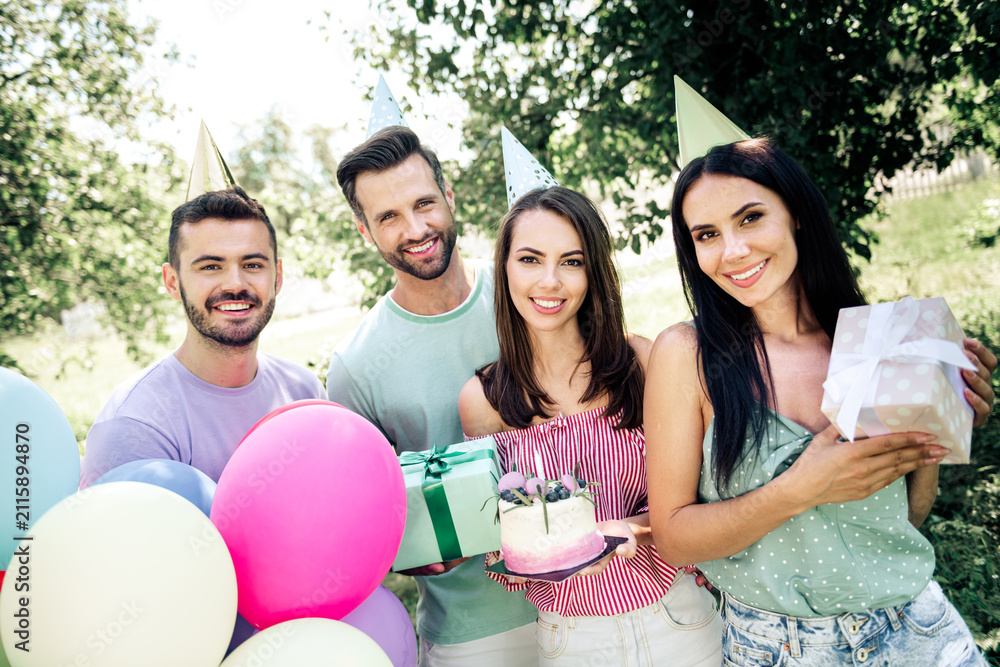 Obraz premium Friends celebrating a birthday party outdoors with cake, balloons and gifts, smiling young adults wearing party hats in a sunny garden