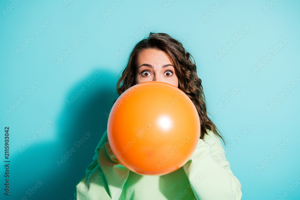 Obraz premium Young woman blowing up a large orange balloon with surprised expression against a turquoise background for playful party and celebration concept