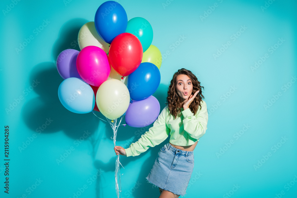 Obraz premium Young woman holding colorful balloons and looking surprised against a turquoise background in a casual denim skirt at a cheerful party celebration