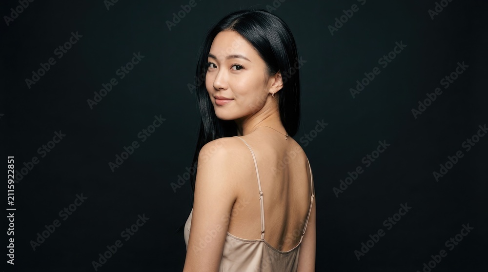 Fototapeta premium 長い黒髪の美しい若いアジア人女性が、黒い背景の前でシルクのドレスを着て優雅にポーズをとっている。