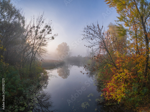 A serene autumn morning reveals a misty river flanked by vibrant colorful trees reflecting softly on the calm water under a hazy blue sky