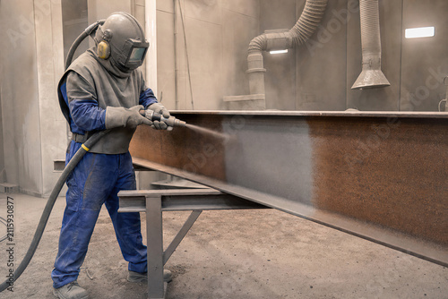 Sandblaster cleans rusty steel beam with abrasive tool. Protective mask and gear worn by worker in factory. Metal surface preparation, industrial maintenance and corrosion removal.