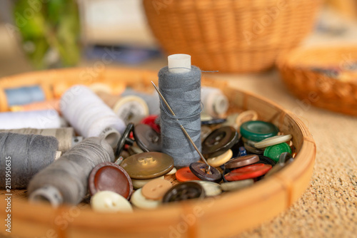 Bild auf Leinwand Close-up of a sewing thread spool with a threaded needle surrounded by assorted buttons and sewing supplies in a wooden tray