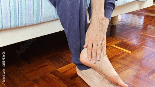 Quadro su tela Elderly man painfully massages his ankle while sitting on bed.