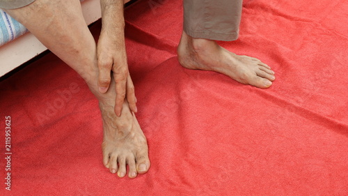 Bild auf Leinwand Elderly man painfully massages his ankle while sitting on bed, with his feet resting on red terry cloth