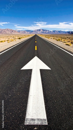 Endless asphalt road with a large white arrow pointing towards distant mountains