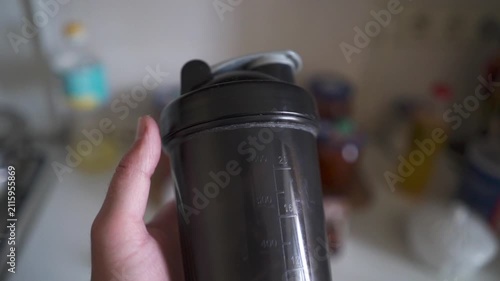 POV shot of person walking through kitchen holding black protein shaker bottle before workout.
