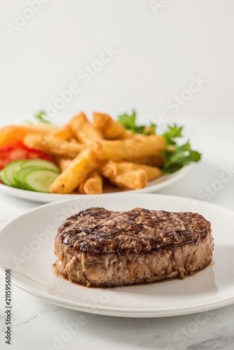 A juicy steak with clear grill marks served on a white plate with French fries and fresh slices of cucumber and tomato.