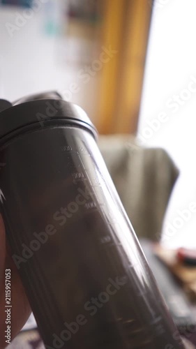 POV silhouette shot of athlete holding fitness shaker bottle walking in room against bright window.