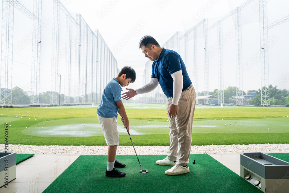 Obraz premium Junior Golf Lesson with a male coach giving posture guidance to an Asian boy holding a club on a practice mat at a spacious outdoor driving range
