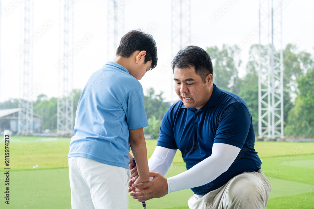 Fototapeta premium Junior Golf Lesson with a male golf coach kneeling to correct an Asian boy's grip and club position during practice at a bright outdoor driving range