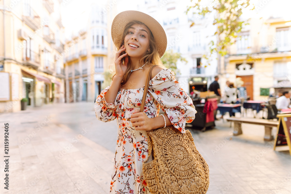 Fototapeta premium Woman walks in city center wearing a dress and hat while holding a woven bag during sunny day