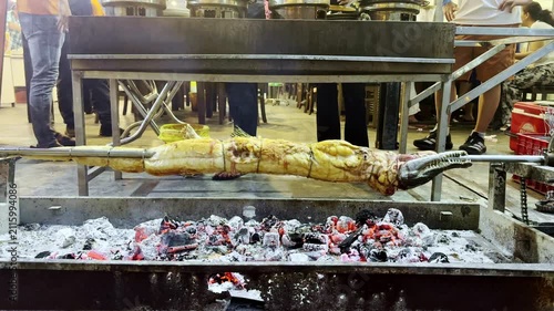 Whole crocodile roasting on a rotating spit over hot charcoal during outdoor barbecue cooking. Traditional exotic cuisine prepared over an open fire.