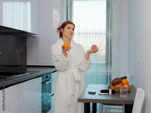 Фотография A woman stands in her kitchen holding an apple and an orange while looking at her glucometer