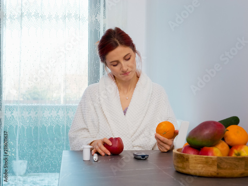 A woman in a kitchen checks her blood sugar level with a glucometer and thinks about eating an apple or an orange. Fruits are on the table along with glucose tools.