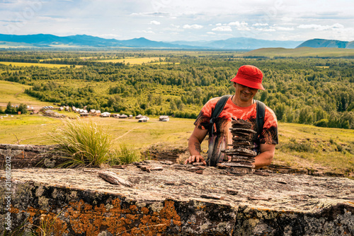 фотографія Man with backpack places tower of flat carne stones on rock outcrop with panoramic view of mountain valley