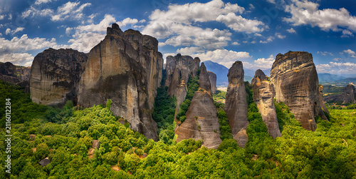 Aerial view of Meteora rock formations surrounded by lush green forests under a bright blue sky with scattered clouds in Trikala, Greece.